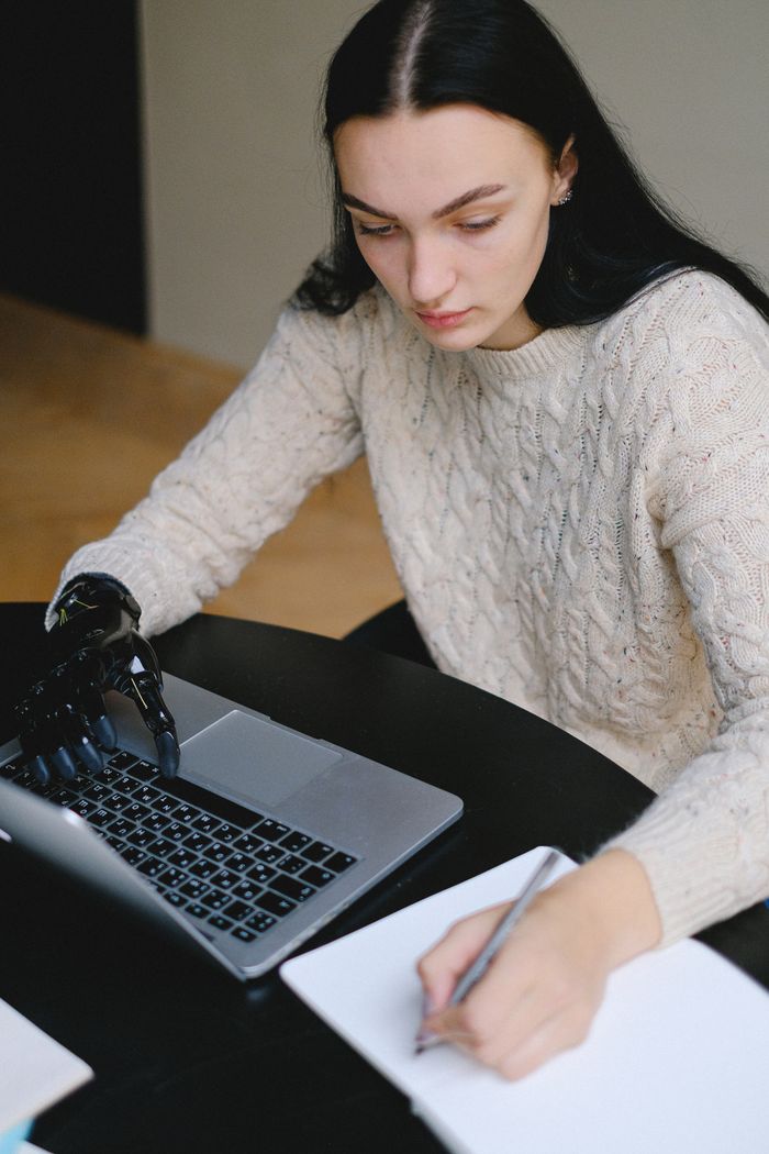 A woman with a prosthetic hand is using a laptop indoors, writing notes on paper.