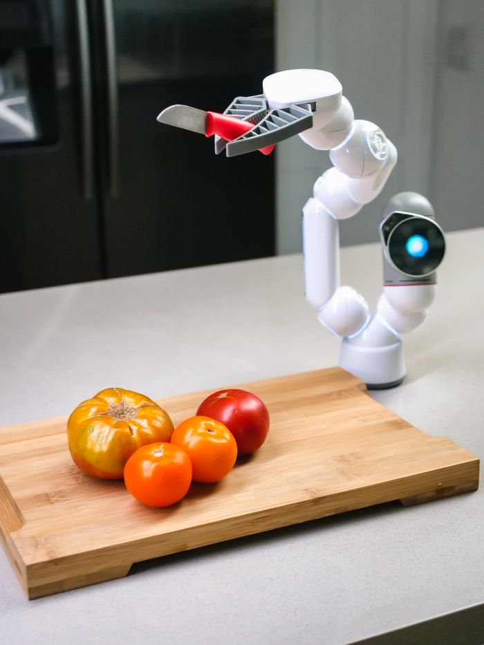 A robotic arm with a knife is slicing tomatoes on a wooden chopping board. Innovative kitchen technology.